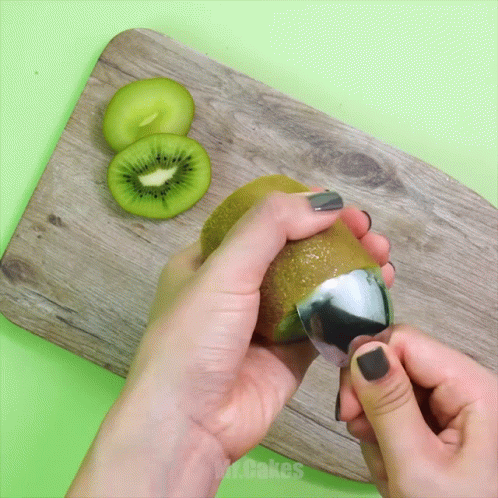 A Person Peeling A Kiwi With A Spoon On A Wooden Cutting Board GIF