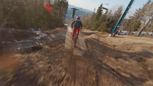 A Person Riding A Bike Down A Dirt Road With A Ski Lift In The Background GIF