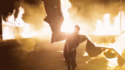 A Person Standing In Front Of A Fire With A Flag Flying In The Air GIF