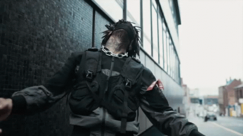 A Person Wearing A Black Jacket And A Mask With A Chain Around Their Neck Is Walking Down The Street GIF
