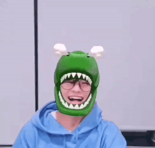 A Person Wearing A Green Dinosaur Hat With A Mouth And Teeth On It . GIF