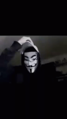 A Person Wearing A Hooded Jacket And A Anonymous Mask Is Waving At The Camera . GIF
