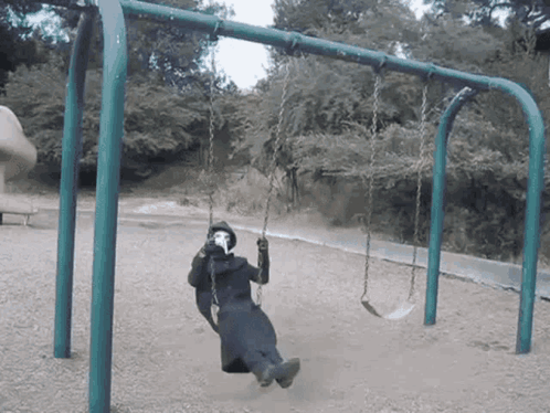 A Person Wearing A Mask Sits On A Swing In A Park GIF