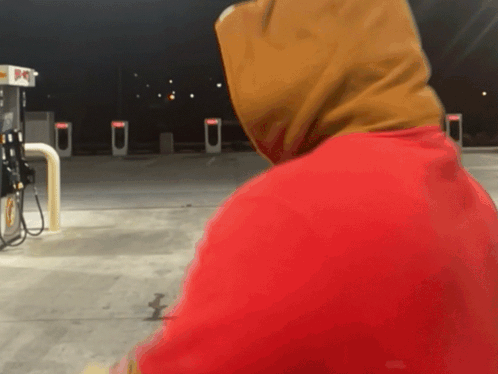 A Person Wearing A Red Hoodie Stands In Front Of A Gas Pump GIF