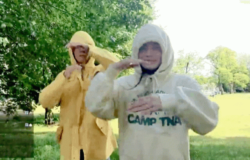 A Person Wearing A White Camp Tna Sweatshirt Stands Next To Another Person Wearing A Yellow Raincoat GIF
