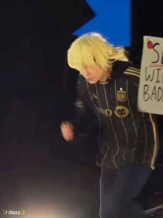 A Person Wearing A Yellow Wig And A Black Jacket With The Letter M On It GIF
