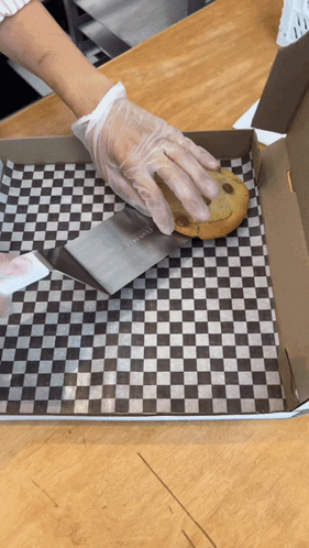 A Person Wearing Gloves Is Cutting A Chocolate Chip Cookie In A Box With A Spatula GIF
