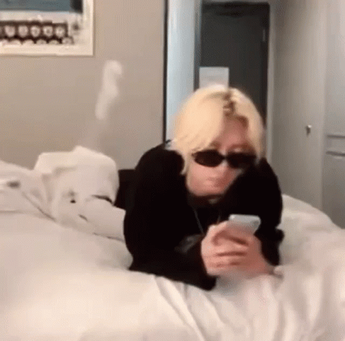 A Person Wearing Sunglasses Is Laying On A Bed Looking At Their Phone . GIF
