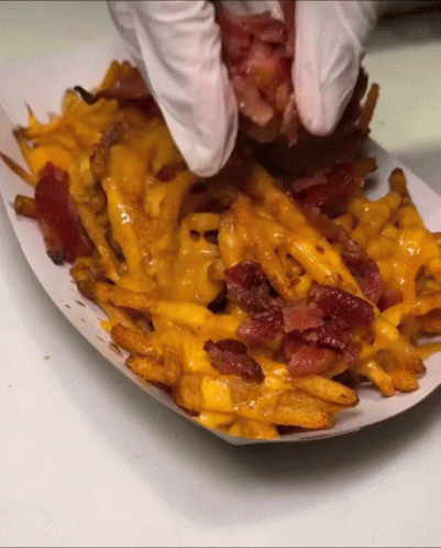A Person Wearing White Gloves Holds A Piece Of Bacon Over French Fries GIF