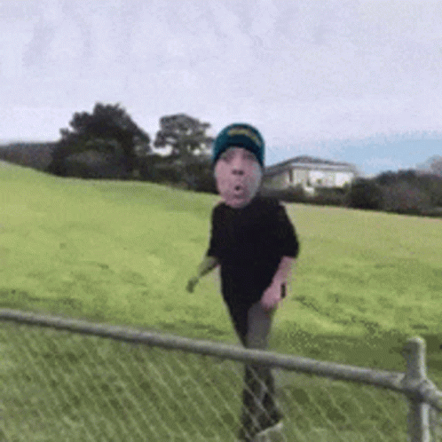 A Person With A Face On Their Head Is Walking Through A Grassy Field Behind A Chain Link Fence . GIF
