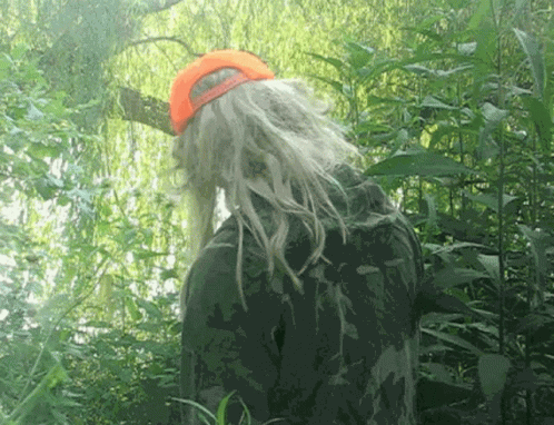 A Person With Long Blonde Hair And An Orange Hat Stands In The Woods GIF