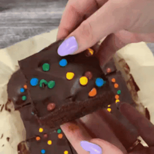 A Person With Purple Nails Is Holding A Piece Of Chocolate Cake With Sprinkles On Top . GIF