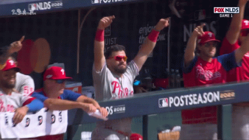 A Philadelphia Phillies Player Stands In A Dugout With His Arms In The Air GIF