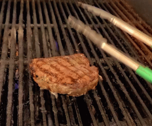 A Piece Of Meat Is Being Cooked On A Grill With Tongs GIF