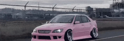 A Pink Car Is Driving Down A Road Next To A Chain Link Fence . GIF
