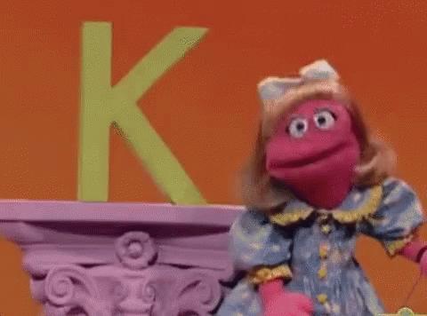 A Pink Puppet Is Standing In Front Of A Green Letter K On A Shelf . GIF