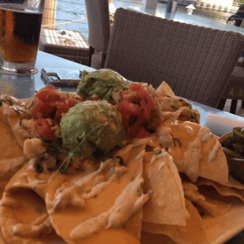 A Plate Of Nachos Sits On A Table With A Glass Of Beer In The Background GIF
