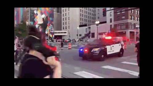 A Police Car Is Driving Down A City Street With A Crowd Behind It . GIF