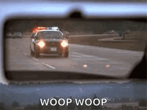 A Police Car Is Driving Down The Road In The Rear View Mirror Of A Car . GIF