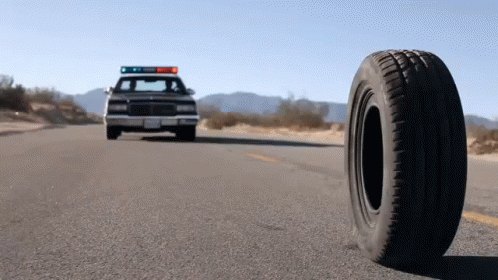 A Police Car Is Driving Down The Road With A Tire On The Side Of The Road GIF