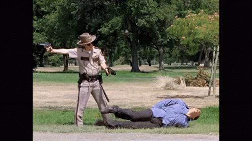 A Police Officer Is Pointing A Gun At A Man Laying On The Grass GIF