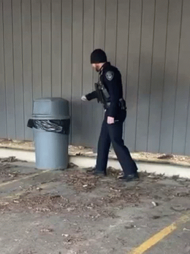 A Police Officer Is Walking Towards A Trash Can . GIF
