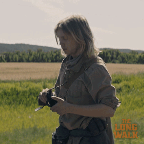 A Poster For The Long Walk Shows A Woman Holding A Camera In A Field GIF