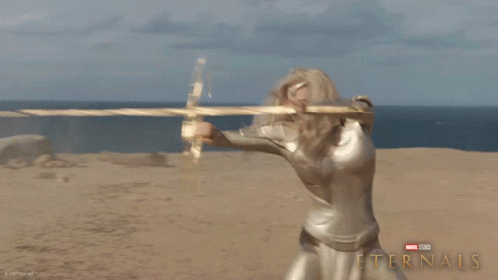 A Poster For The Movie Eternals Shows A Blonde Woman Holding A Golden Object GIF