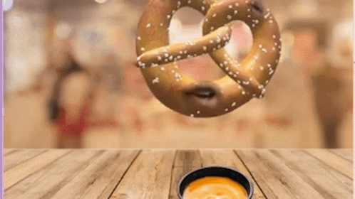 A Pretzel Is Falling Into A Bowl Of Cheese Sauce On A Wooden Table GIF