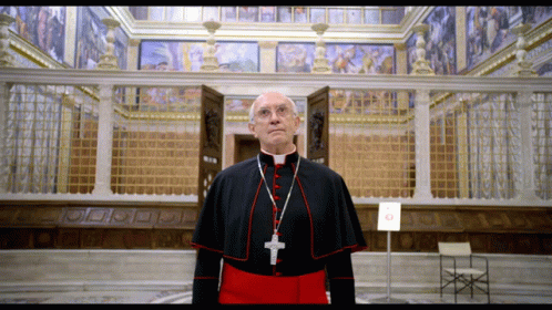 A Priest With A Cross Around His Neck Stands In Front Of A Building GIF