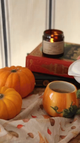 A Pumpkin Shaped Cup Sits On A Table Next To A Candle And Books GIF