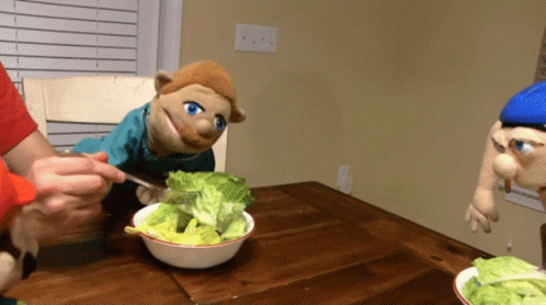 A Puppet Is Eating Lettuce From A Bowl On A Table GIF