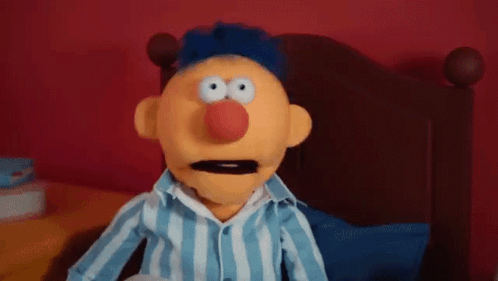 A Puppet Is Sitting On A Bed Wearing A Blue And White Striped Shirt And A Red Nose . GIF