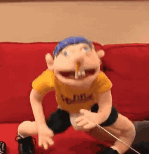A Puppet Is Sitting On A Red Couch With A Stick In His Mouth . GIF