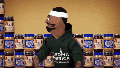 A Puppet Wearing A Green Shirt That Says Feeding America Stands In Front Of Jars Of Peanut Butter GIF