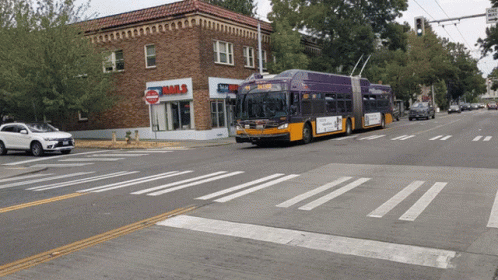 A Purple Bus Is Driving Down The Street In Front Of A Building That Says Nails GIF
