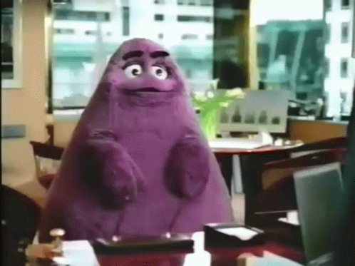 A Purple Cartoon Character Is Sitting At A Table In An Office GIF