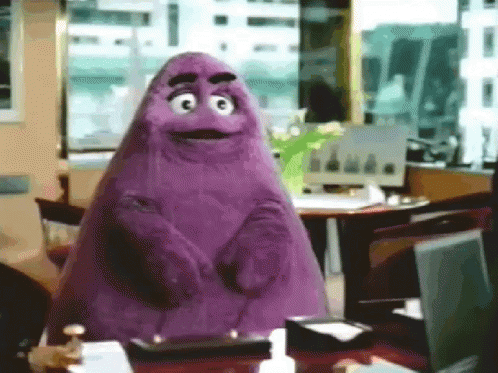 A Purple Cartoon Character Is Standing In Front Of A Desk With A Laptop On It . GIF