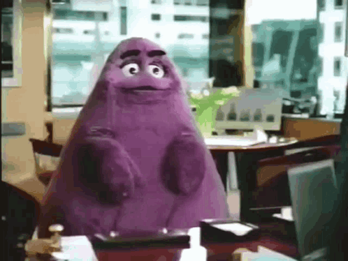 A Purple Mcdonald 'S Mascot Is Sitting At A Desk In An Office . GIF