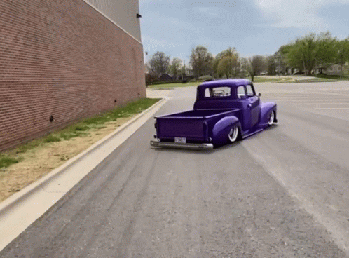A Purple Truck Is Driving Down A Street With A Brick Building In The Background And A Few Trees GIF