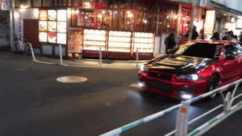 A Red Car Is Driving Down A Street In Front Of A Restaurant With A Sign That Says ' Noodles ' On It GIF