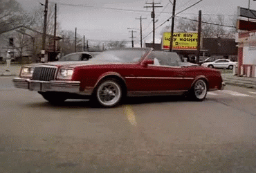 A Red Car Is Driving Down A Street In Front Of A Sign That Says We Buy Ugly Houses GIF