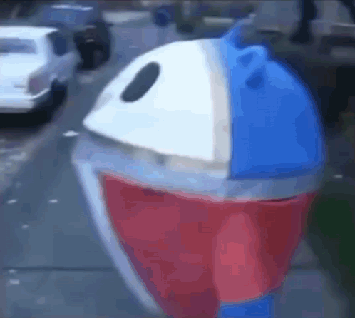 A Red , White And Blue Trash Can Is Sitting On The Side Of A Street . GIF