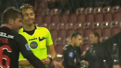A Referee Wearing A Yellow Shirt With The Word Diadora On It Is Standing On A Soccer Field . GIF