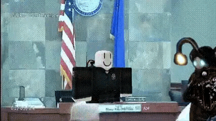 A Roblox Character Is Sitting At A Desk In Front Of A Computer In A Courtroom With An American Flag In The Background . GIF