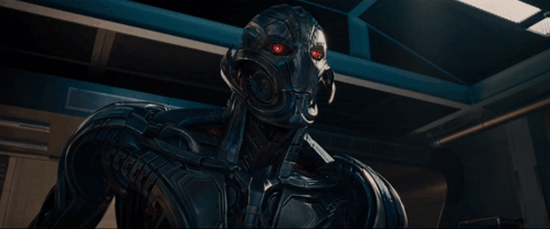 A Robot Is Standing In A Dark Room With A Blue Light Behind Him GIF
