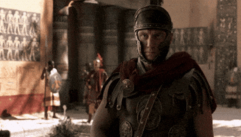 A Roman Soldier With A Helmet On Stands In Front Of A Wall With Images Of People On It GIF