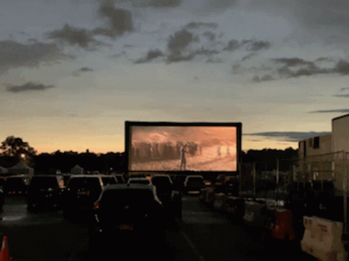 A Row Of Cars Are Parked In Front Of A Large Screen That Shows A Movie GIF