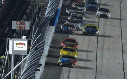 A Row Of Race Cars Are Driving Down A Track With A Sign That Says Cook Out GIF