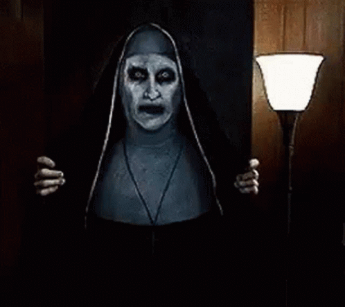 A Scary Nun Is Standing In A Dark Room Holding A Lamp And Looking At The Camera . GIF